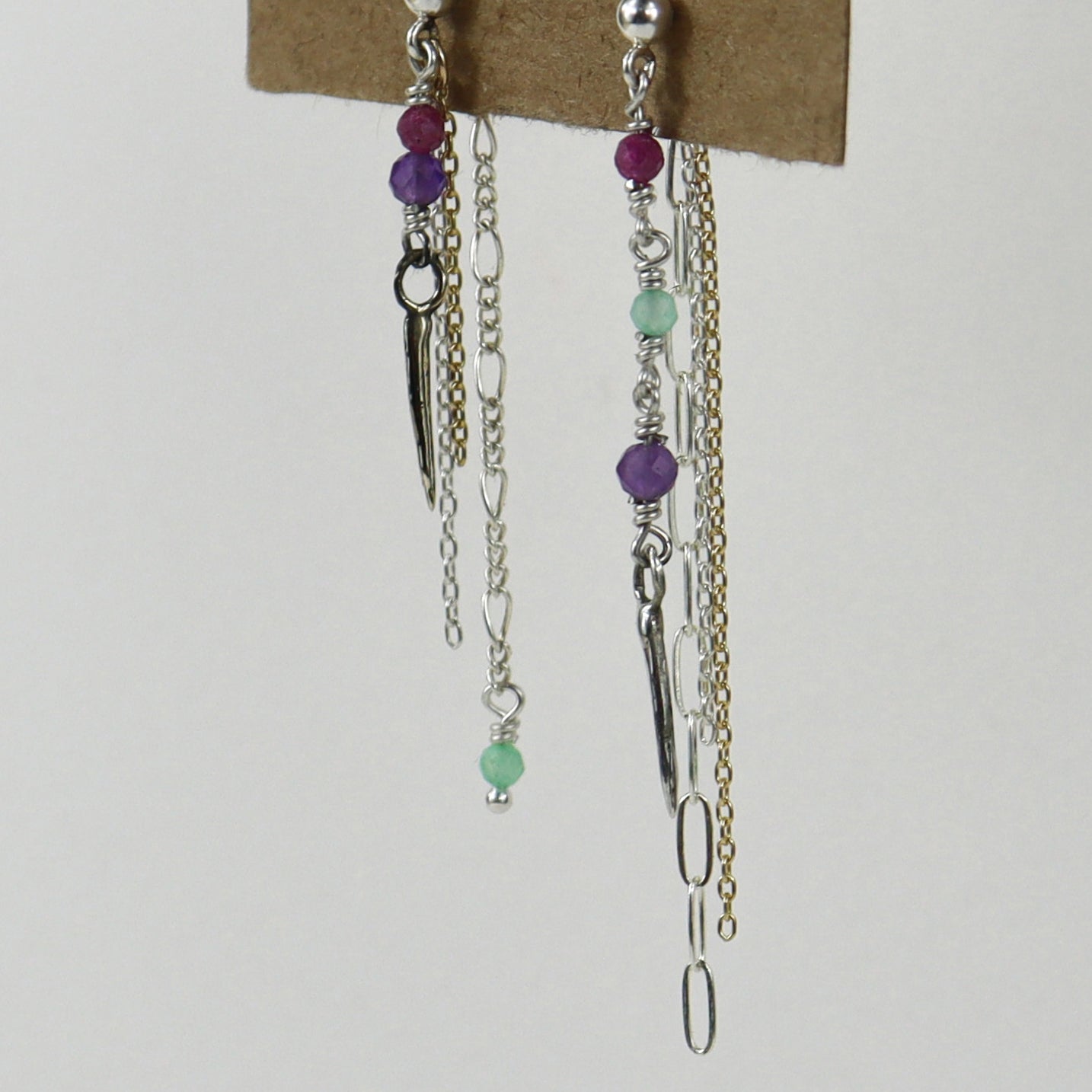 A pair of silver-toned dangle earrings with purple and mint green beads, handmade by Kria World, displayed on a brown kraft card against a light gray background.