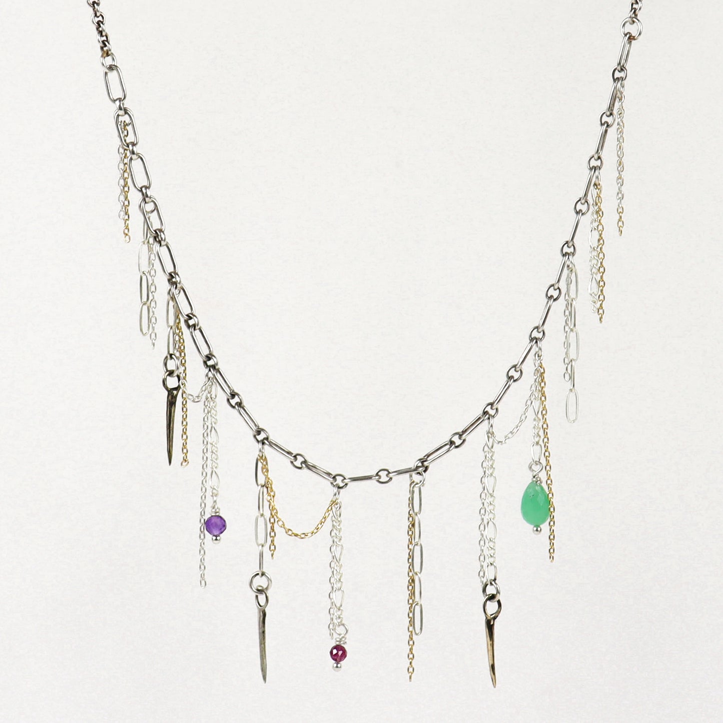 A silver-toned layered necklace handmade by Kria World with multiple dangling chains, small metallic charms, and three colorful gemstone accents in purple, red, and green, displayed against a plain off-white background.