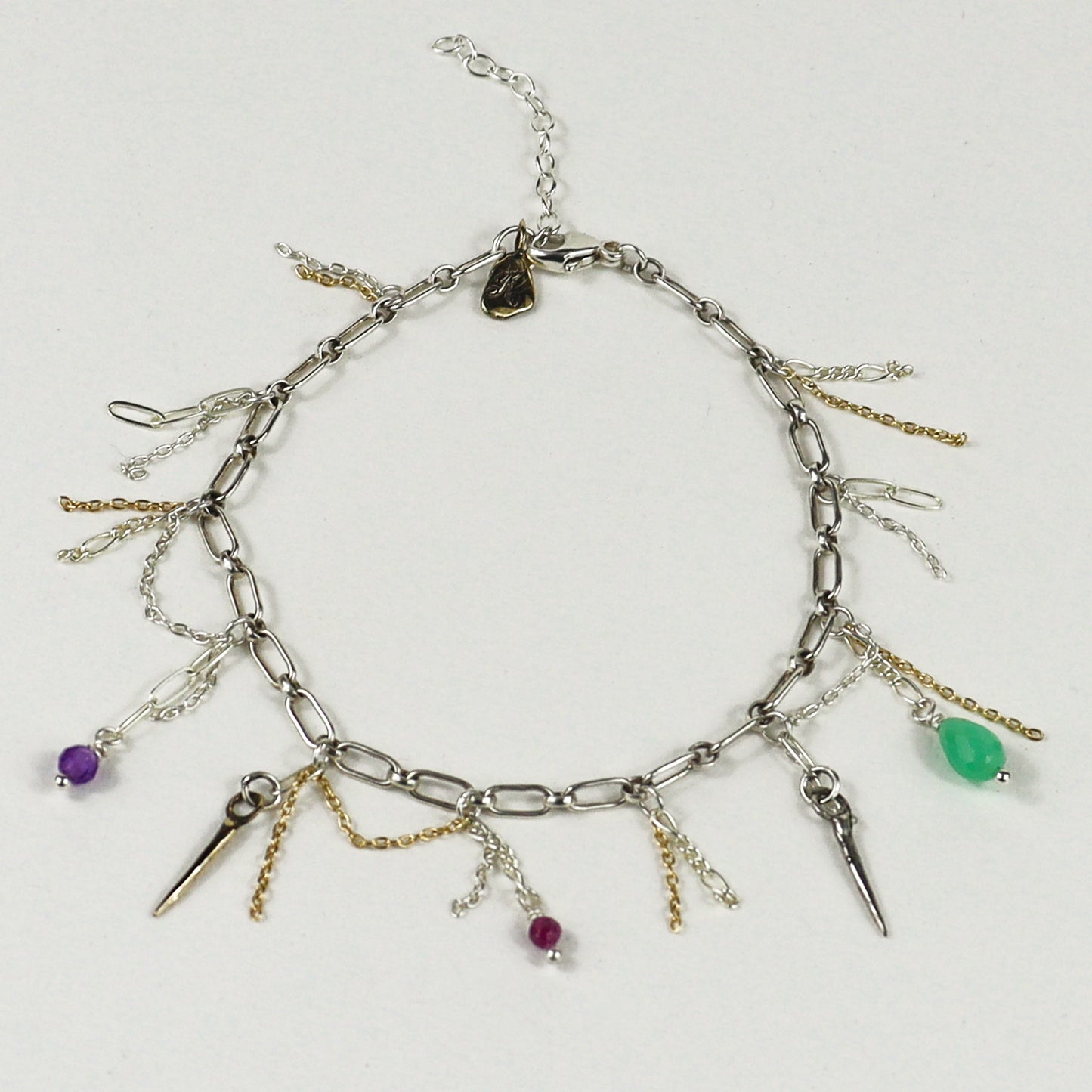 A silver chain bracelet handmade by Kria World with multiple dangling charms, including spikes, chains, and small beads in purple, green, and magenta, displayed on a light gray background.