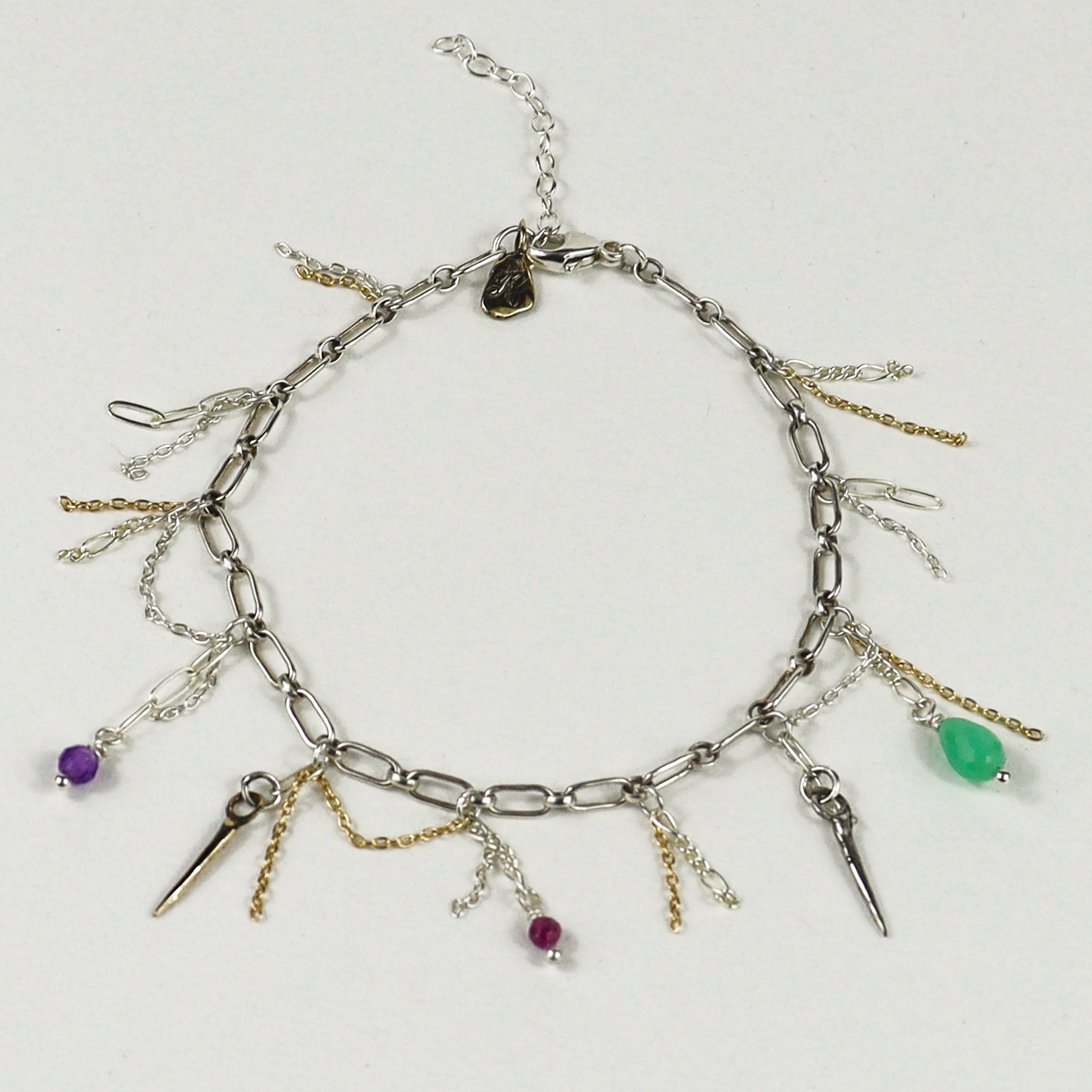 A silver chain bracelet handmade by Kria World with multiple dangling charms, including spikes, chains, and small beads in purple, green, and magenta, displayed on a light gray background.
