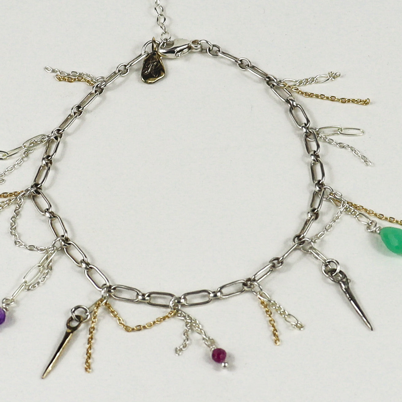 A silver chain bracelet handmade by Kria World with multiple dangling charms, including spikes, chains, and small beads in purple, green, and magenta, displayed on a light gray background.