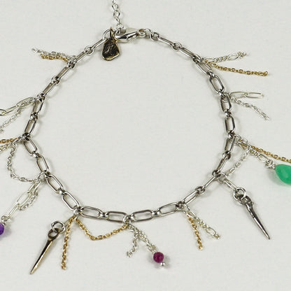 A silver chain bracelet handmade by Kria World with multiple dangling charms, including spikes, chains, and small beads in purple, green, and magenta, displayed on a light gray background.