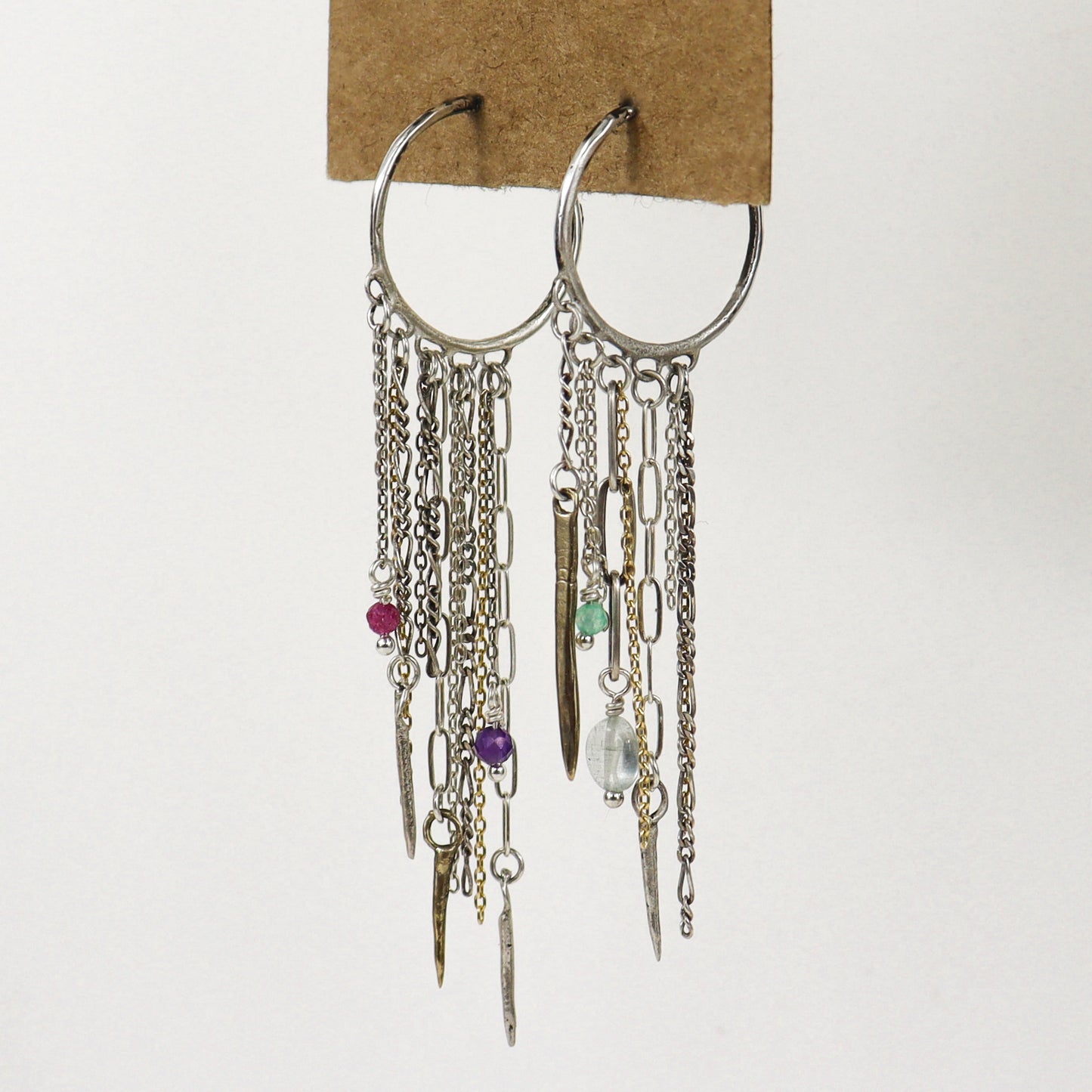 A pair of silver hoop earrings with multiple dangling chains, links, beads, and small charms, including a pink bead, purple bead, green bead, and clear faceted bead, handmade by Kria World and displayed on a brown cardboard backing against a white background.