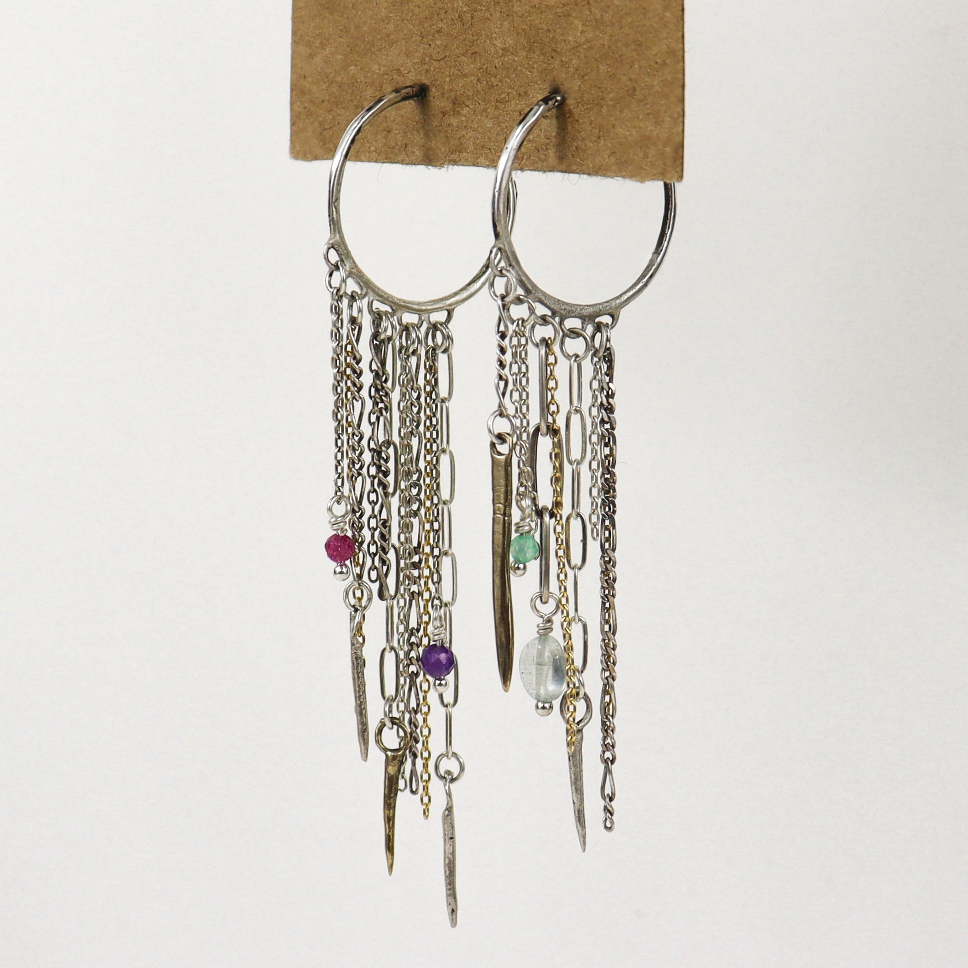 A pair of silver hoop earrings with multiple dangling chains, links, beads, and small charms, including a pink bead, purple bead, green bead, and clear faceted bead, handmade by Kria World and displayed on a brown cardboard backing against a white background.