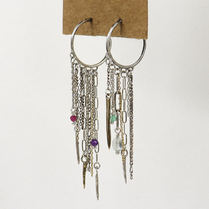A pair of silver hoop earrings with multiple dangling chains, links, beads, and small charms, including a pink bead, purple bead, green bead, and clear faceted bead, handmade by Kria World and displayed on a brown cardboard backing against a white background.