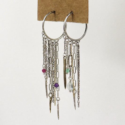 A pair of silver hoop earrings with multiple dangling chains, links, beads, and small charms, including a pink bead, purple bead, green bead, and clear faceted bead, handmade by Kria World and displayed on a brown cardboard backing against a white background.