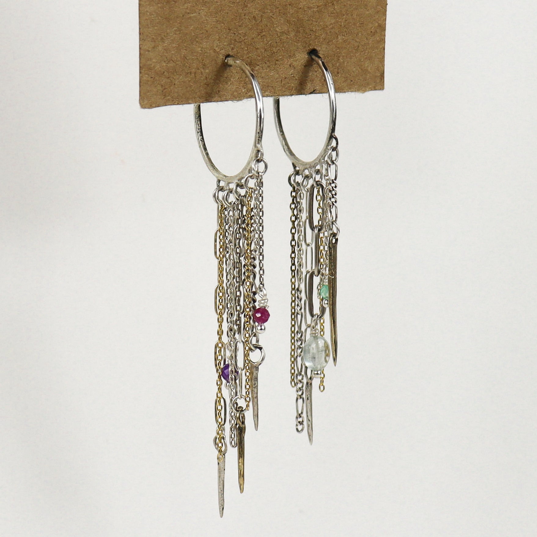 A pair of silver hoop earrings with multiple dangling chains, links, beads, and small charms, including a pink bead, purple bead, green bead, and clear faceted bead, handmade by Kria World and displayed on a brown cardboard backing against a white background.