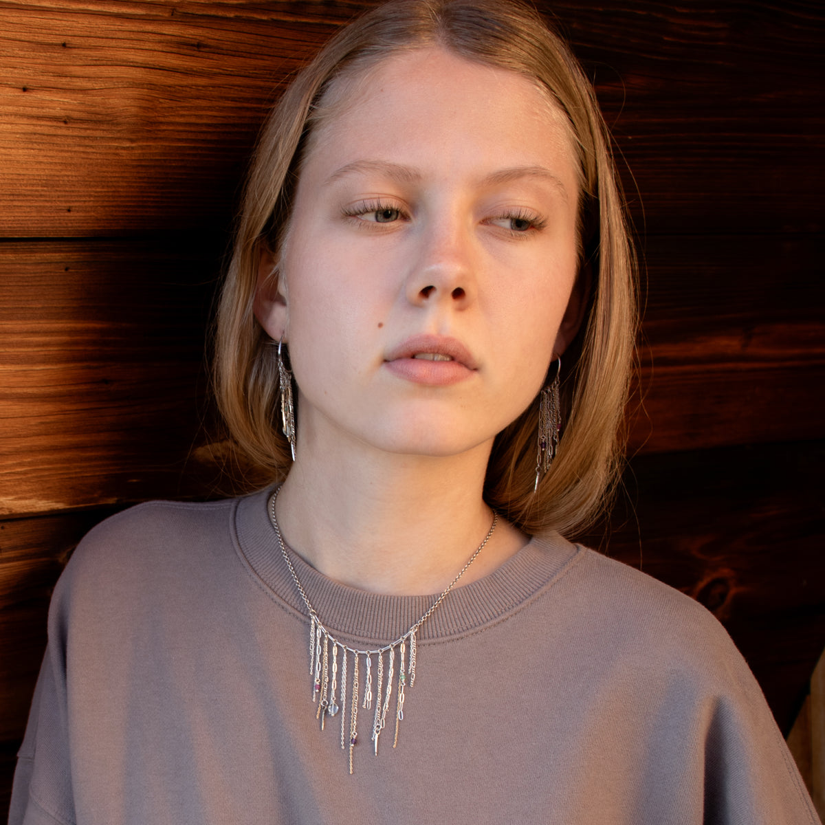 A woman with blonde hair wearing a silver fringe necklace and matching drop earrings by Kria World set against a dark wood-paneled background.