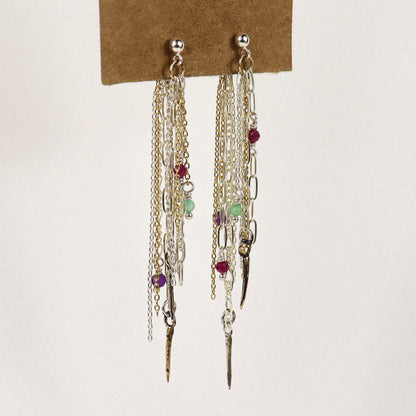 A pair of long dangling earrings displayed on a brown card against a plain light background. The earrings handmade by Kria World feature multiple layers of silver and gold-tone chains, small metallic charms, and several tiny faceted beads in red, green, and purple.