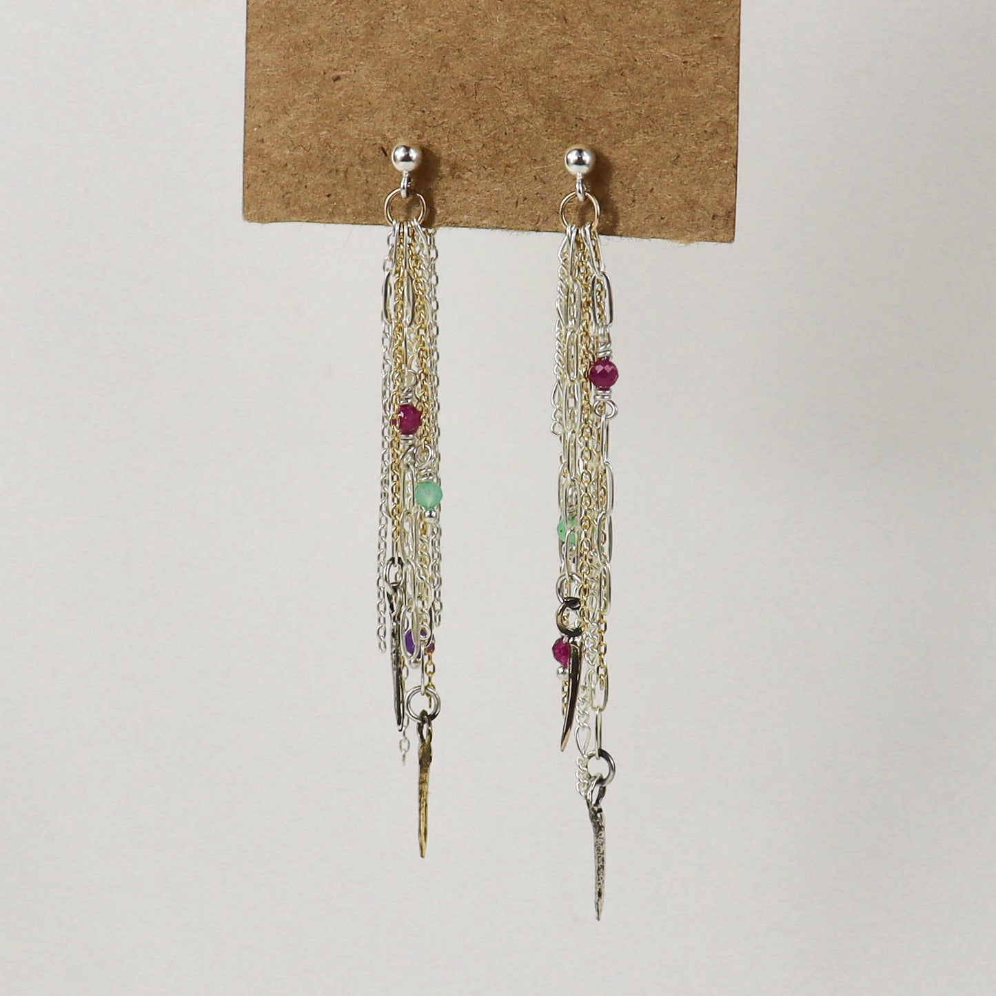 A pair of long dangling earrings displayed on a brown card against a plain light background. The earrings handmade by Kria World feature multiple layers of silver and gold-tone chains, small metallic charms, and several tiny faceted beads in red, green, and purple.