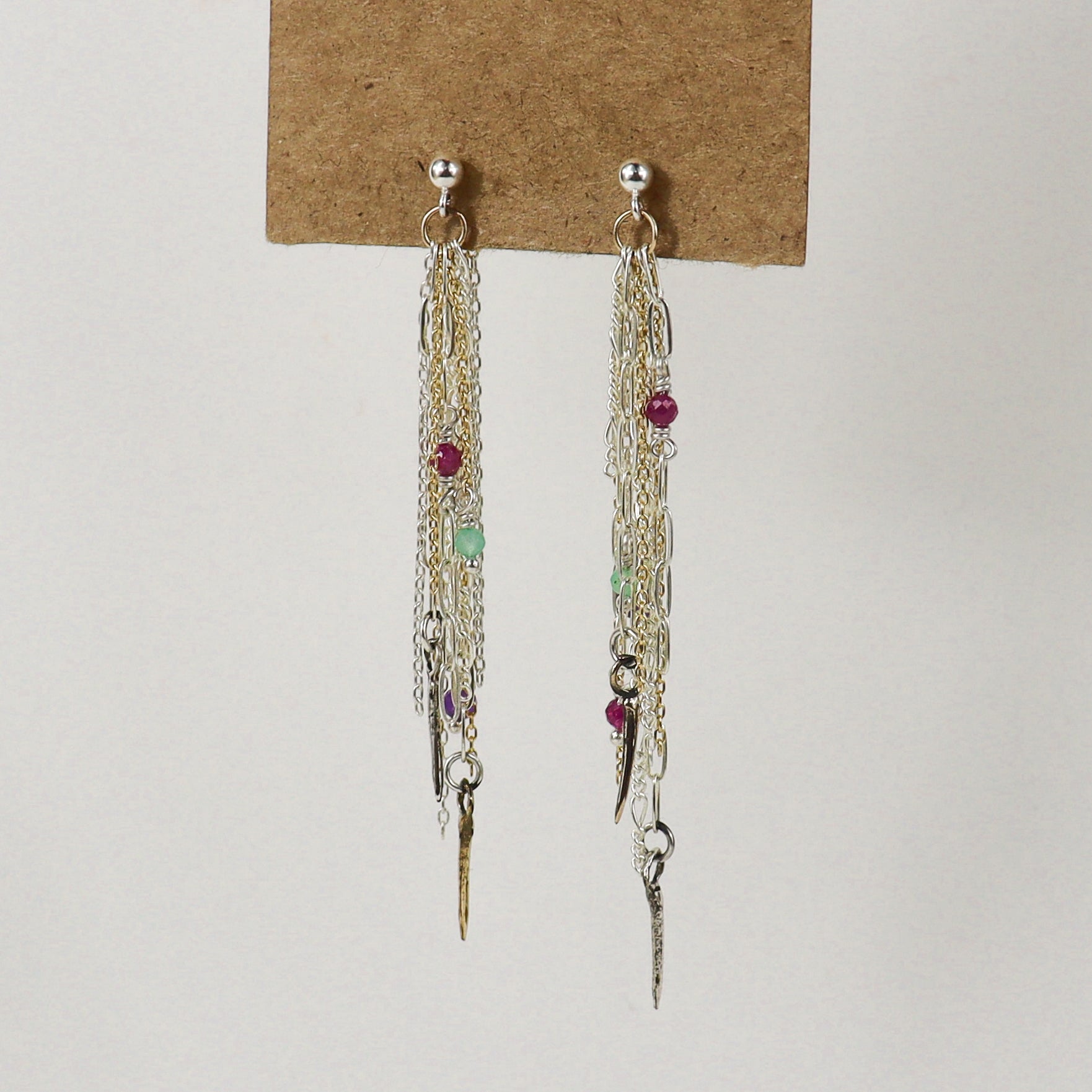 A pair of long dangling earrings displayed on a brown card against a plain light background. The earrings handmade by Kria World feature multiple layers of silver and gold-tone chains, small metallic charms, and several tiny faceted beads in red, green, and purple.