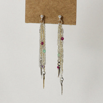A pair of long dangling earrings displayed on a brown card against a plain light background. The earrings handmade by Kria World feature multiple layers of silver and gold-tone chains, small metallic charms, and several tiny faceted beads in red, green, and purple.