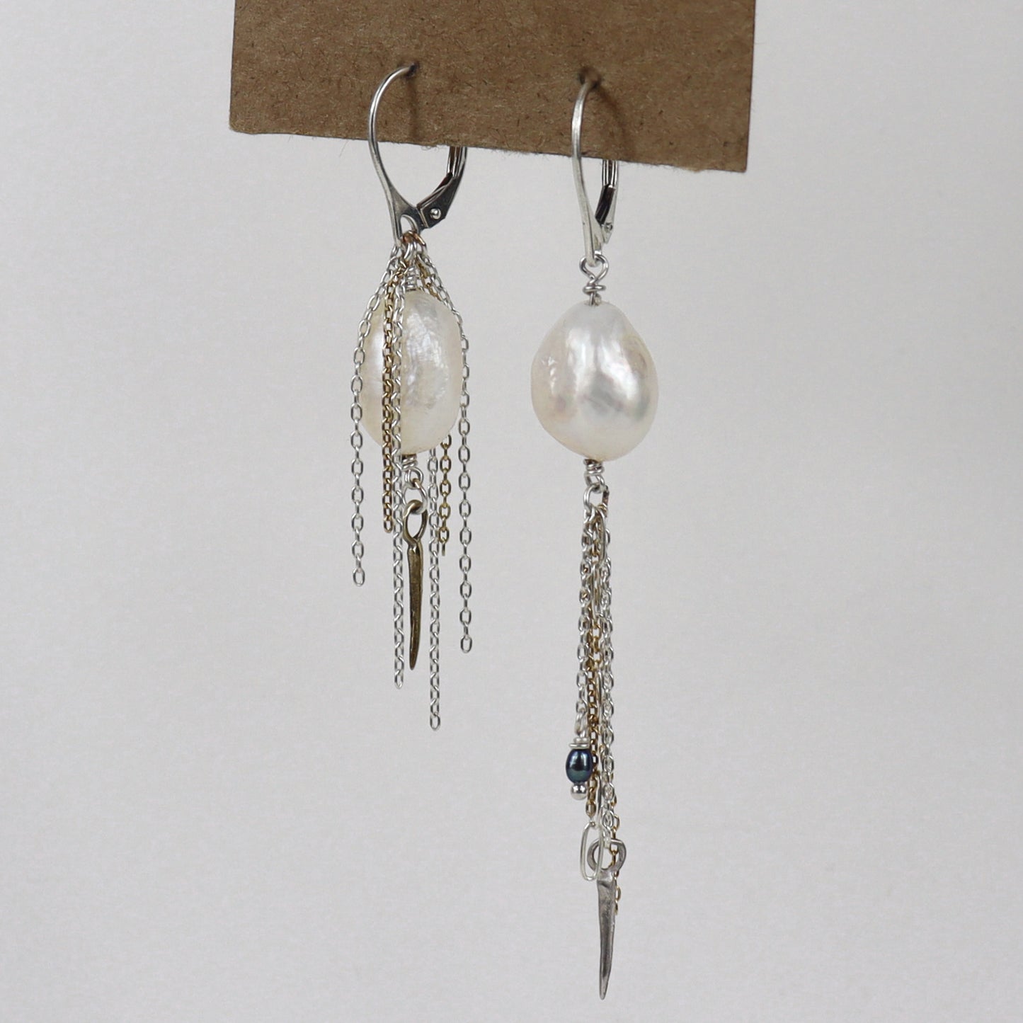 Asymmetrical sterling silver hoop earrings with two large freshwater pearls and one tiny black pearl with gold-filled and silver double mixed-chain tassels 2 and 3 inches each shown on paper tag on white background.