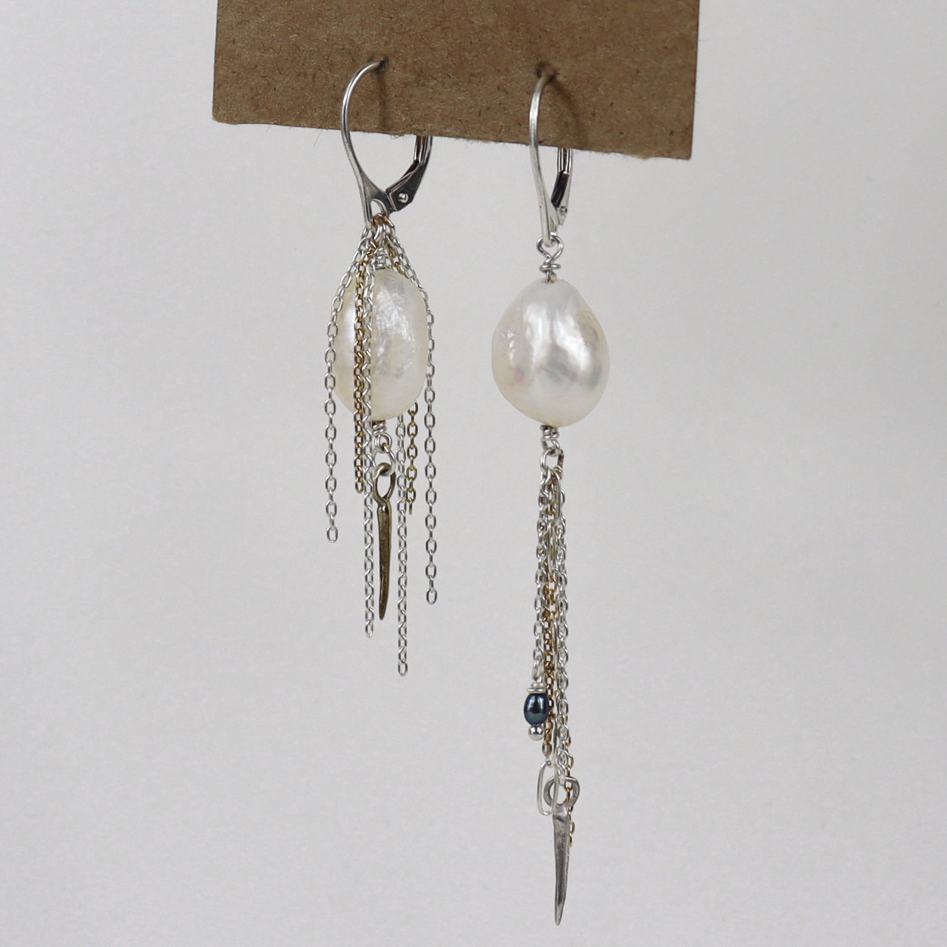 Asymmetrical sterling silver hoop earrings with two large freshwater pearls and one tiny black pearl with gold-filled and silver double mixed-chain tassels 2 and 3 inches each shown on paper tag on white background.