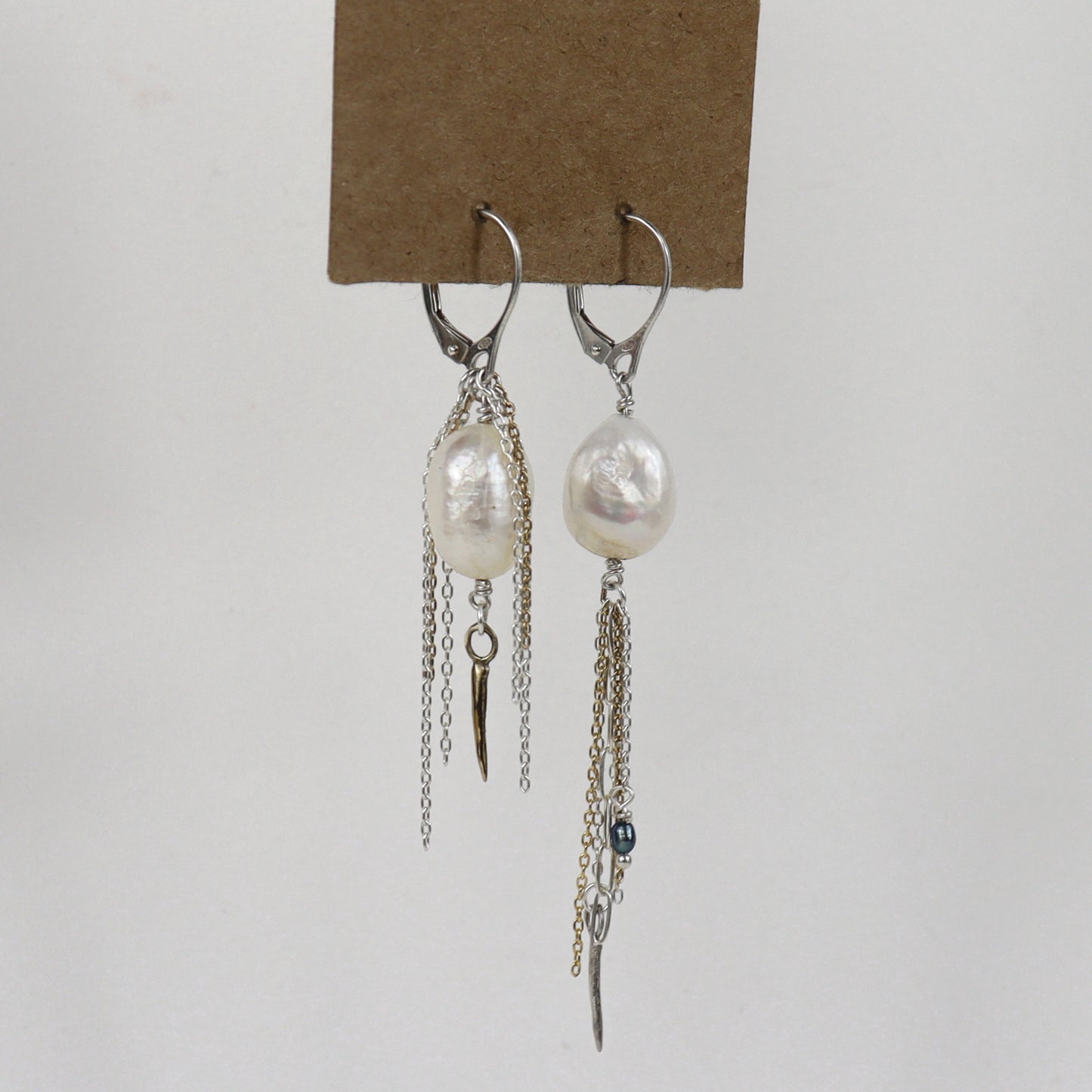 Asymmetrical sterling silver hoop earrings with two large freshwater pearls and one tiny black pearl with gold-filled and silver double mixed-chain tassels 2 and 3 inches each shown on paper tag on white background.
