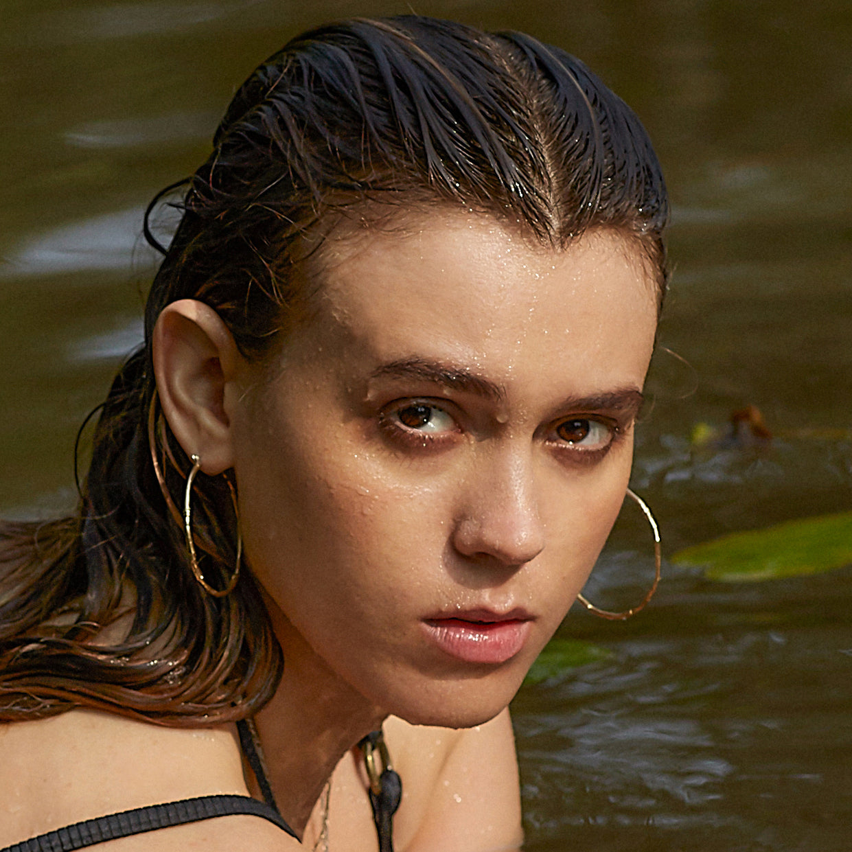 Woman with wet hair standing in water, wearing gold hoop earrings from Kria World jewelry.