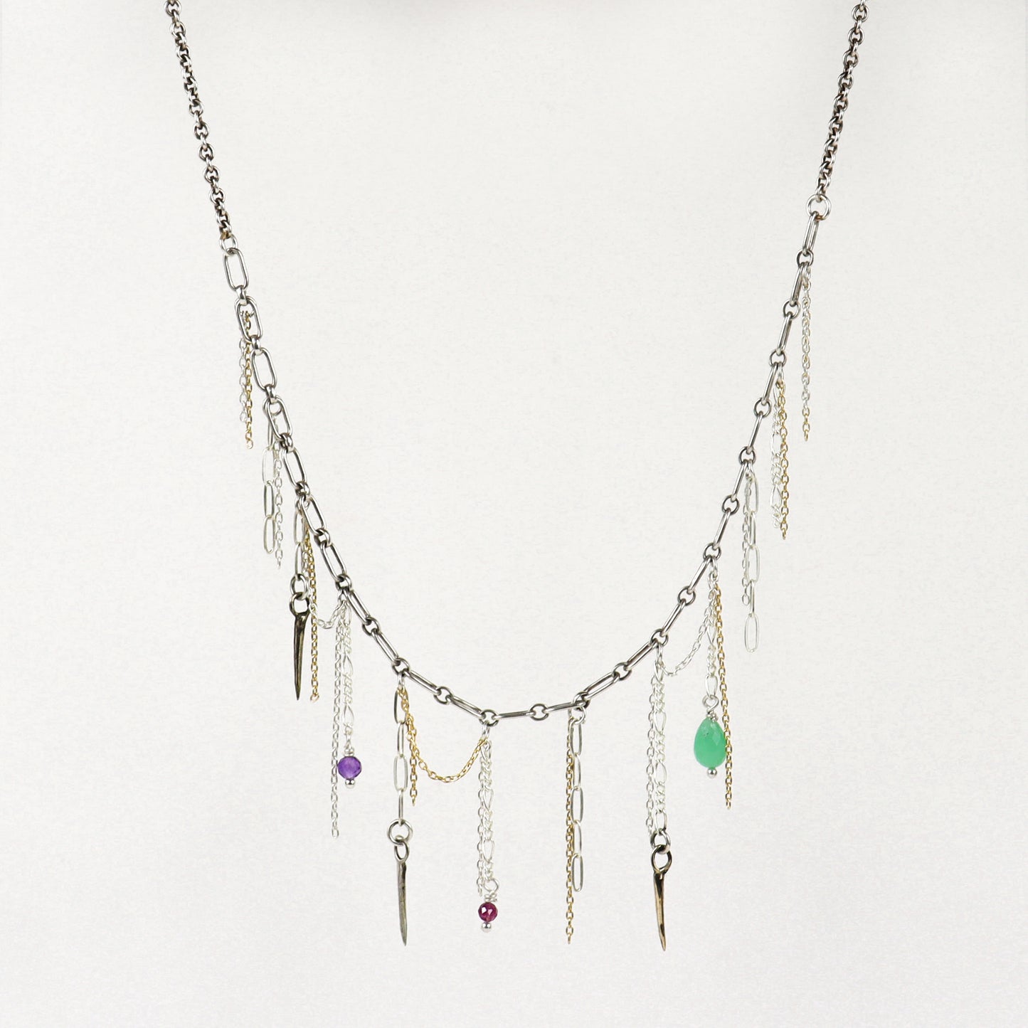 A silver-toned layered necklace handmade by Kria World with multiple dangling chains, small metallic charms, and three colorful gemstone accents in purple, red, and green, displayed against a plain off-white background.