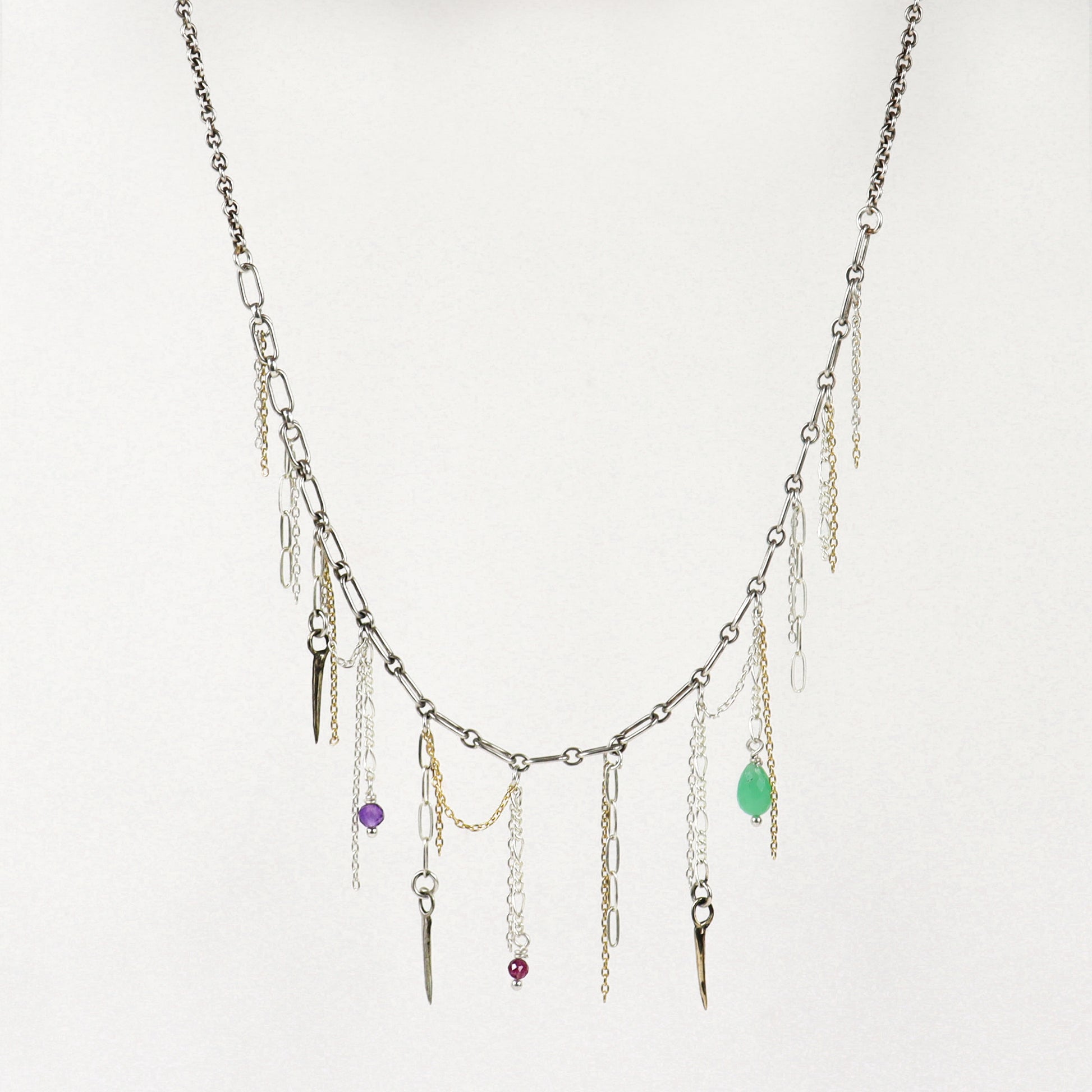 A silver-toned layered necklace handmade by Kria World with multiple dangling chains, small metallic charms, and three colorful gemstone accents in purple, red, and green, displayed against a plain off-white background.