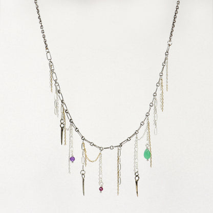 A silver-toned layered necklace handmade by Kria World with multiple dangling chains, small metallic charms, and three colorful gemstone accents in purple, red, and green, displayed against a plain off-white background.