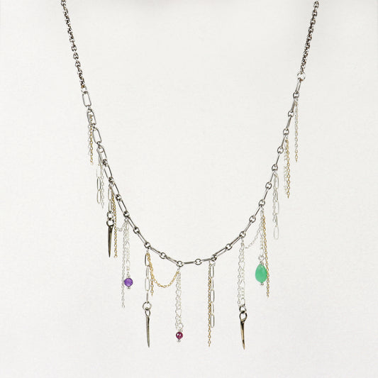 A silver-toned layered necklace handmade by Kria World with multiple dangling chains, small metallic charms, and three colorful gemstone accents in purple, red, and green, displayed against a plain off-white background.