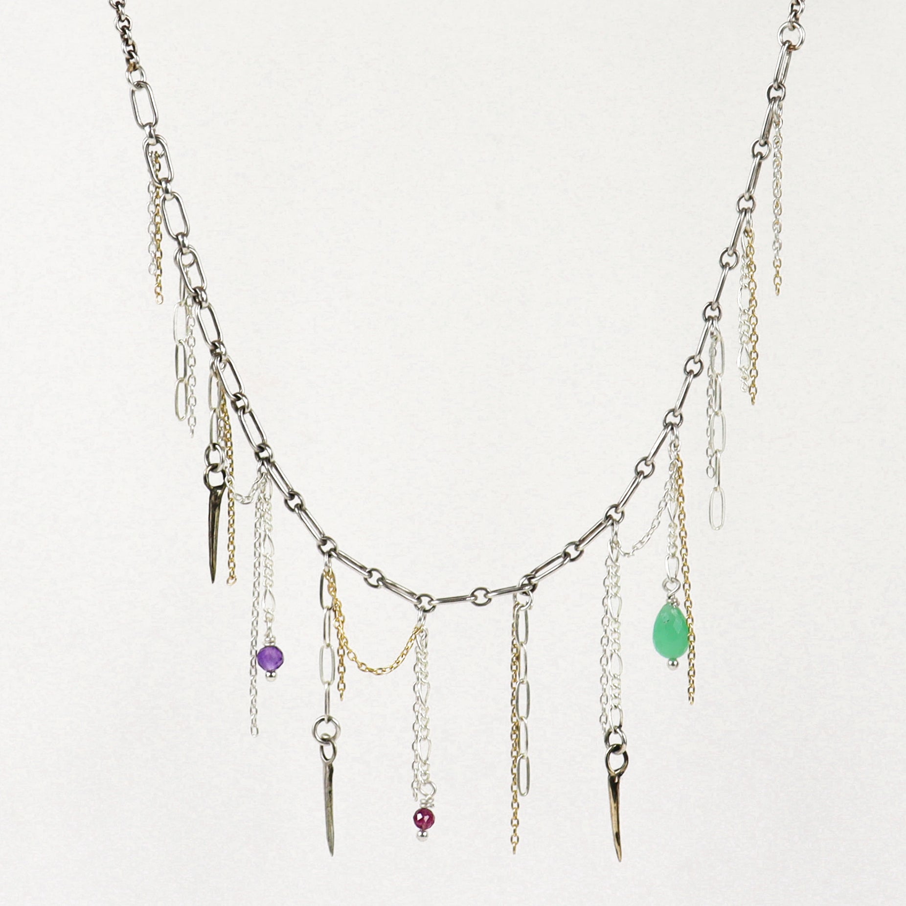 A silver-toned layered necklace handmade by Kria World with multiple dangling chains, small metallic charms, and three colorful gemstone accents in purple, red, and green, displayed against a plain off-white background.