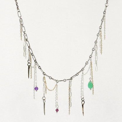 A silver-toned layered necklace handmade by Kria World with multiple dangling chains, small metallic charms, and three colorful gemstone accents in purple, red, and green, displayed against a plain off-white background.