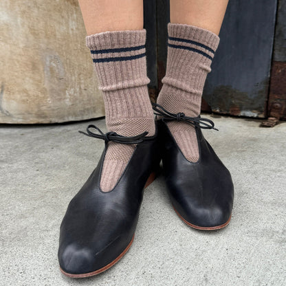 A person wearing cocoa ribbed crew socks by Le Bon Shoppe with navy stripes and black leather Mary Jane shoes, standing on a concrete floor.