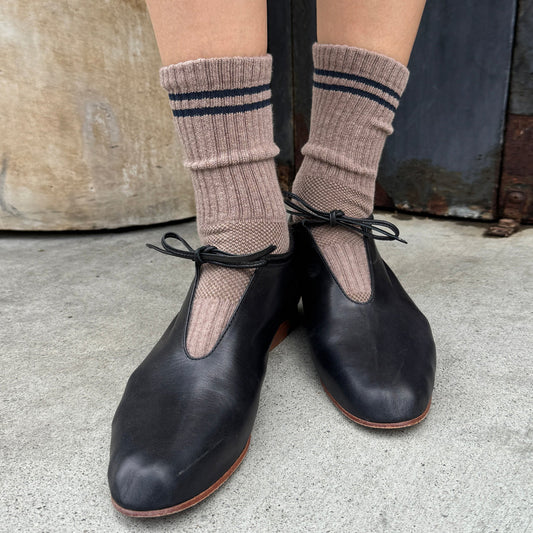 A person wearing cocoa ribbed crew socks by Le Bon Shoppe with navy stripes and black leather Mary Jane shoes, standing on a concrete floor.