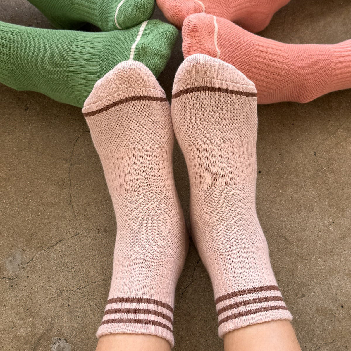 Three pairs of knit crew socks in green, Bellini, and peach y Le Bon Shoppe are arranged on a concrete floor, with the front pink socks being worn on feet.