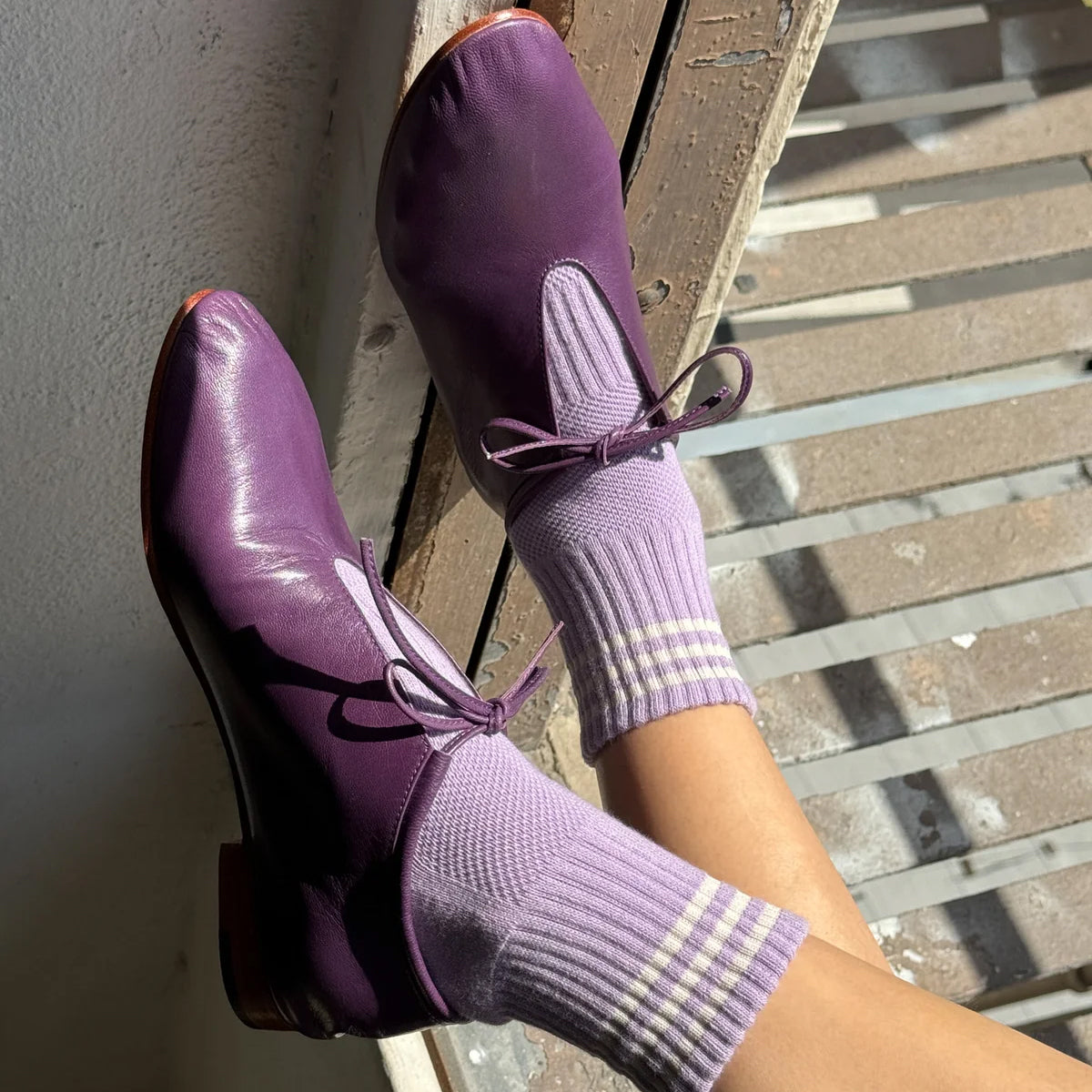 Purple leather Mary Jane-style shoes with ribbon laces, worn with light purple socks by Le Bon Shoppe, shown on feet against a rustic wooden slat background.